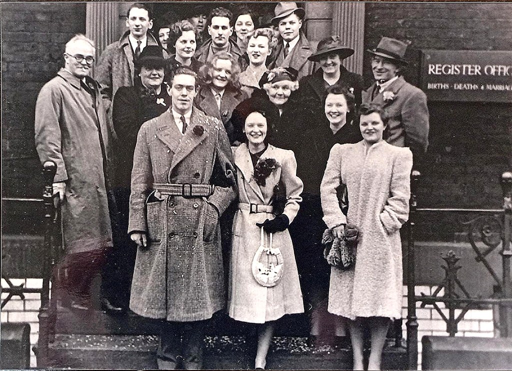 A group of more than a dozen smartly dressed people standing on the steps outside a registry office after the marriage of Denys Fisher and Betty. Denys and Betty are in the middle at the front and are linking arms.